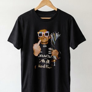 Puede incluir: Una camiseta negra de manga corta con un estampado gr&aacute;fico de una persona con gafas blancas de Las Vegas, sosteniendo un trofeo y haciendo un gesto con el dedo coraz&oacute;n. La persona viste un traje de carreras negro con logotipos de Oracle y Red Bull.