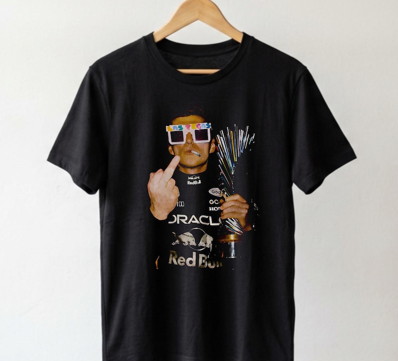 Puede incluir: Una camiseta negra de manga corta con un estampado gr&aacute;fico de una persona con gafas blancas de Las Vegas, sosteniendo un trofeo y haciendo un gesto con el dedo coraz&oacute;n. La persona viste un traje de carreras negro con logotipos de Oracle y Red Bull.