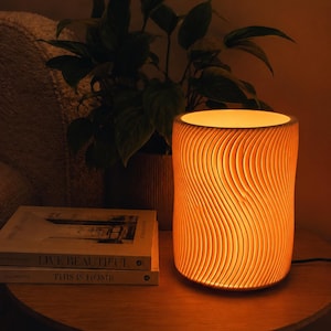 May include: A cylindrical table lamp emitting a warm, orange light. The lamp features a textured, wave-like design. It rests on a wooden table beside a stack of books and a potted plant. The books are titled "Live Beautiful" and "This is Home".