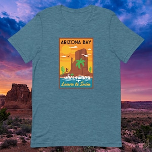 May include: Teal t-shirt featuring a graphic with the text "Arizona Bay" and "Learn to Swim." The graphic depicts a desert landscape with a sun, palm tree, and rock formations. The shirt is made of a soft material.