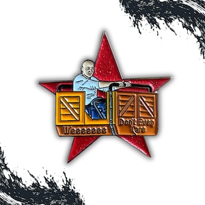 May include: A red star-shaped pin featuring a bald man in a light blue shirt and blue jeans, operating a cart with the words "Weeeeeeee" and "Don't Even Care". The cart is made of wooden crates.