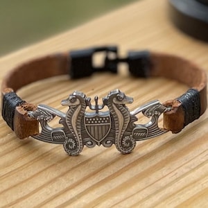 May include: A brown leather bracelet with a silver metal charm featuring two seahorses flanking a shield with the American flag. The bracelet has a black braided cord and a silver metal clasp.