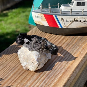 May include: A black braided bracelet with a silver charm featuring two seahorses flanking a shield with the US Coast Guard emblem.