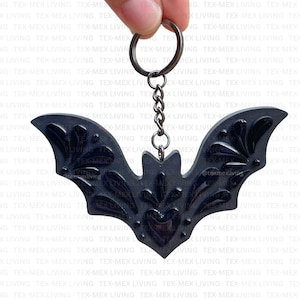 May include: A black bat-shaped keychain featuring an embossed floral pattern on the wings and a central heart-shaped detail. The bat is attached to a dark metal split ring by a short chain, creating a cohesive dark-toned accessory for keys.