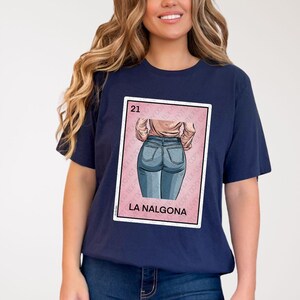 Op de afbeelding: Een marineblauw t-shirt met een roze en witte illustratie van een vrouw in jeans. De illustratie is in de stijl van een traditionele Mexicaanse speelkaart. De tekst "21" en "LA NALGONA" staat op de kaart gedrukt.