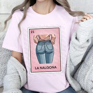 Op de afbeelding: Een roze T-shirt met een afbeelding van een vrouw in blauwe jeans. De afbeelding is een parodie op een traditionele Mexicaanse speelkaart, met het nummer 21 en de tekst "La NalgonA".