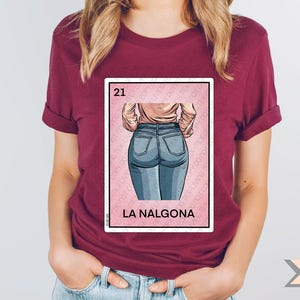 Op de afbeelding: Een maroon t-shirt met een afbeelding van de rug van een vrouw in blauwe jeans. De afbeelding is in de stijl van een traditionele Mexicaanse speelkaart met het nummer 21 en de tekst "La Nalgona".