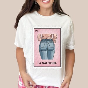 Op de afbeelding: Wit T-shirt met een roze en witte illustratie van een vrouw in blauwe jeans. De illustratie is in de stijl van een traditionele Mexicaanse speelkaart. De tekst "21" en "LA NALGONA" staat op de kaart gedrukt.