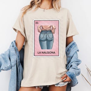 Op de afbeelding: Een beige T-shirt met een afbeelding van een roze kaart met het nummer 21 en de tekst "LA NALGO&Nu;&Alpha;". De kaart toont een cartoonillustratie van de rug van een vrouw die een blauwe jeans draagt.
