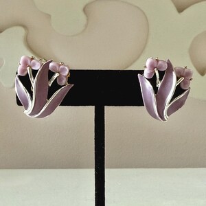 May include: A pair of clip-on earrings featuring a floral design with light purple enamel petals and small, rounded light pink flower buds. The metal edges are silver-toned, outlining the curved leaf shapes and the cluster of three small buds.
