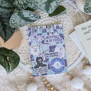 K&ouml;nnte beinhalten: Ein blaues und violettes Notizbuch mit verschiedenen Aufklebern. Die Aufkleber zeigen einen Wolf, eine Zaubertrankflasche, ein Schwert, eine Wolke und ein Zitat, das "Mentally I'm in Yvela" lautet.