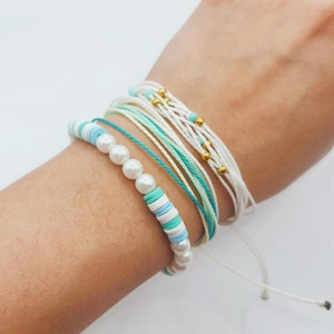 以下が含まれることがあります： 白、青、緑の色合いのブレスレットのコレクション。1つのブレスレットは白い真珠と青と緑のディスクビーズが特徴です。他のブレスレットは紐でできており、一部は金色のビーズが付いています。