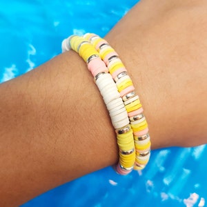 May include: Two beaded bracelets stacked on a wrist. The bracelets feature a mix of pastel colors including yellow, pink, and white, separated by silver-colored spacers. The background is a bright blue.