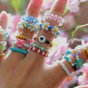 Puede incluir: Una mano muestra una colección de anillos de cuentas en varios colores y diseños. Los anillos presentan patrones florales, de corazón y geométricos, con colores como blanco, azul, rosa y dorado. Los anillos están hechos de pequeñas cuentas.