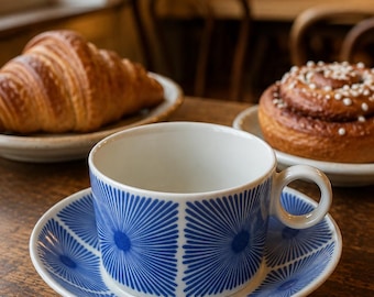 May include: A white teacup and saucer set with a blue geometric pattern, alongside a croissant and a cinnamon roll on a wooden table. The cup and saucer have a radial design. The pastries are on small white plates.