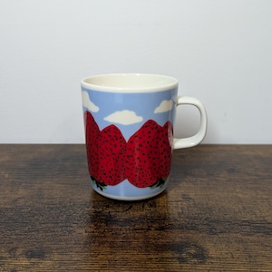 Peut inclure: Mug en céramique blanche avec une anse. Le mug présente un motif de grosses fraises rouges sur un fond bleu clair avec des nuages blancs. Le mug est posé sur une surface en bois.