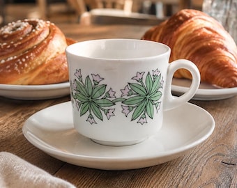 May include: A white teacup and saucer set with a green and pink floral design, alongside a cinnamon roll and a croissant on white plates. The scene is set on a wooden table in a cafe, with a blurred background.