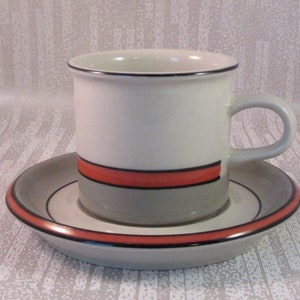 Peut inclure: Un ensemble tasse et soucoupe à café en céramique blanche. La tasse a une base grise, une bande rouge et un bord noir. La soucoupe a un design assorti avec une bande rouge et un bord noir. Idéal pour le café ou le thé.