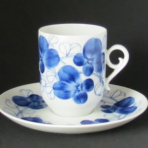 May include: A white porcelain coffee cup and saucer set with a blue floral design. The cup has a unique handle and the saucer features a matching pattern.