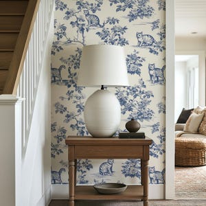 May include: A wooden side table with a white lamp and a blue and white cat-themed wallpaper. The wallpaper features a floral and cat design. A small, round, brown vase and a stack of books sit on the table.