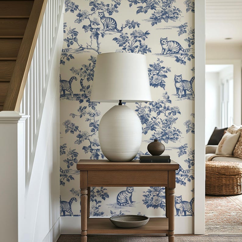 May include: A wooden side table with a white lamp and a blue and white cat-themed wallpaper. The wallpaper features a floral and cat design. A small, round, brown vase and a stack of books sit on the table.
