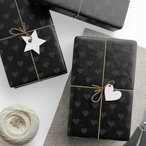 May include: Three black gift boxes wrapped in paper with a pattern of small black hearts. The boxes are tied with twine and decorated with white paper stars and hearts.
