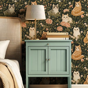 May include: A bedroom scene featuring a light green bedside cabinet with a lamp and books. The wallpaper is a dark green floral pattern with cat illustrations in various poses and colors. The bed has a white pillow and a beige blanket.