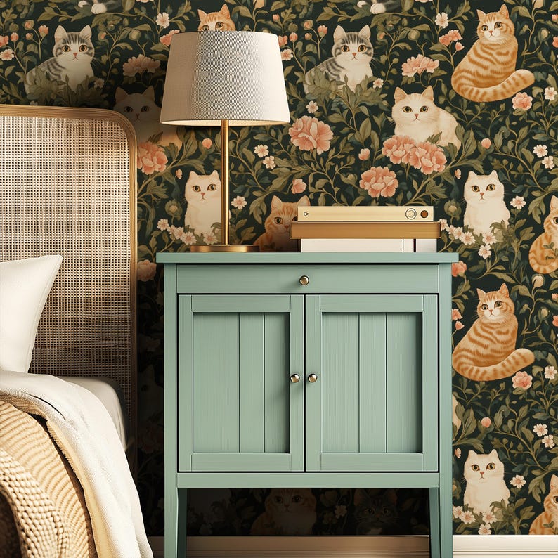 May include: A bedroom scene featuring a light green bedside cabinet with a lamp and books. The wallpaper is a dark green floral pattern with cat illustrations in various poses and colors. The bed has a white pillow and a beige blanket.