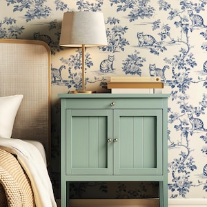 May include: A bedroom scene featuring a light green bedside cabinet with a lamp and books on top. The background is a wallpaper with a blue floral and cat design. A bed with a woven headboard and white bedding is partially visible.