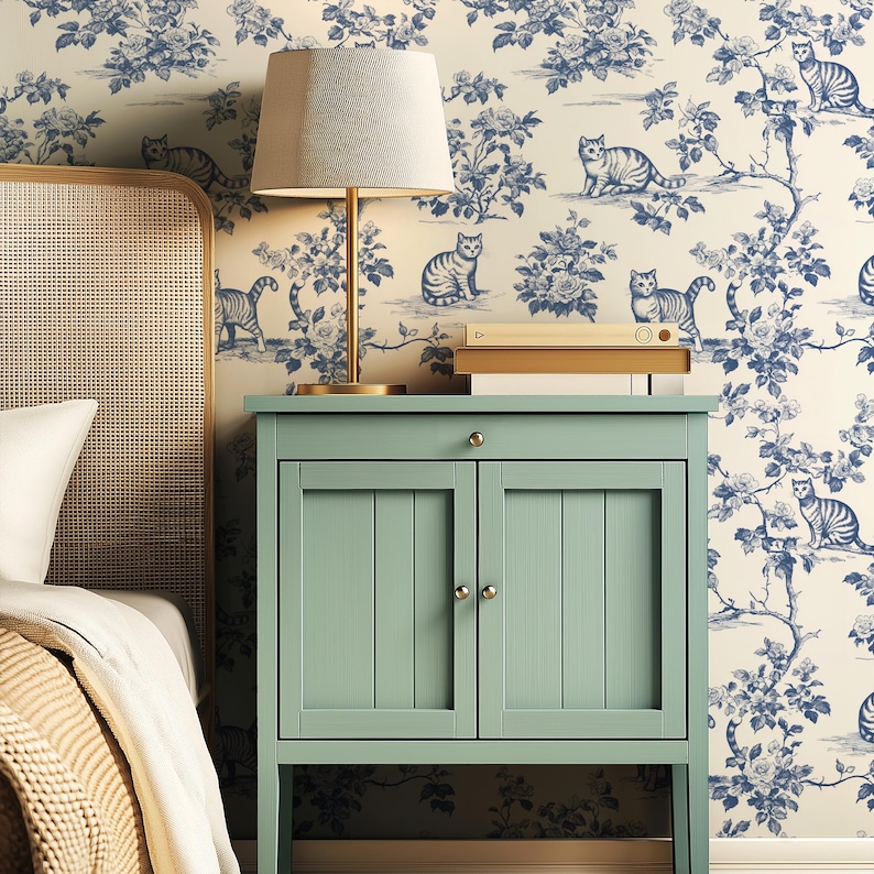May include: A bedroom scene featuring a light green bedside cabinet with a lamp and books on top. The background is a wallpaper with a blue floral and cat design. A bed with a woven headboard and white bedding is partially visible.
