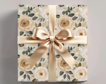 Peut inclure: Une boîte cadeau carrée enveloppée dans du papier à motif de roses beiges et blanches avec des feuilles vert foncé. La boîte est nouée avec un ruban de satin couleur champagne lisse formant un nœud centré sur le devant de l'emballage.