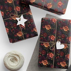 Pode incluir: Tr&ecirc;s pacotes retangulares embrulhados em papel preto com um padr&atilde;o floral de rosas laranja, vermelhas e roxo-escuras. Cada pacote &eacute; amarrado com barbante marrom e adornado com uma etiqueta de papel branco em forma de estrela ou cora&ccedil;&atilde;o.