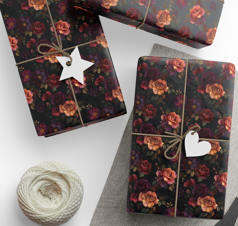 Pode incluir: Tr&ecirc;s pacotes retangulares embrulhados em papel preto com um padr&atilde;o floral de rosas laranja, vermelhas e roxo-escuras. Cada pacote &eacute; amarrado com barbante marrom e adornado com uma etiqueta de papel branco em forma de estrela ou cora&ccedil;&atilde;o.