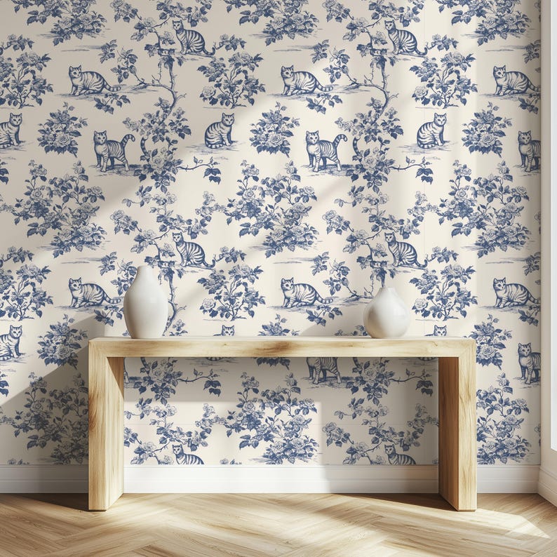May include: Wallpaper featuring a repeating pattern of blue cats and floral elements on a cream background. The design includes various cat poses among stylized flowers and branches. A wooden table with two white vases sits in front of the wallpaper.