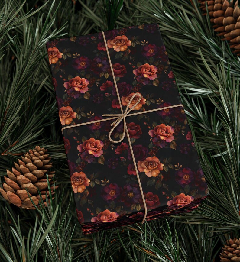 Pode incluir: Um presente retangular embrulhado em papel preto com um padr&atilde;o repetitivo de rosas laranja, vermelhas e roxo-escuras com folhas verdes. O pacote est&aacute; atado com um fio fino castanho-claro disposto em cruz e preso com um la&ccedil;o.