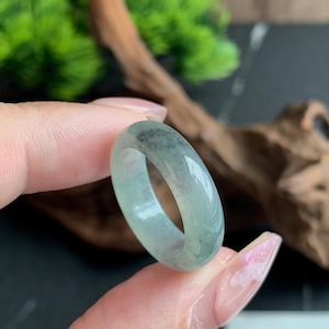 May include: A smooth, polished band ring crafted from translucent light green jadeite stone. The ring features natural mottled dark green inclusions throughout the polished surface, creating a unique pattern within the solid, circular stone band.
