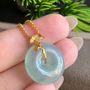 May include: A circular, translucent pale green jade pendant suspended from a thin gold beaded chain. The pendant features a gold ginkgo leaf accent attached to the top, which connects the circular stone to the delicate gold chain.