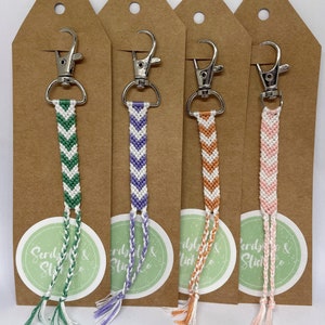 May include: Four colourful friendship bracelet keychains with white chevron patterns. The keychains are attached to brown card stock tags with the text "Scribble & Stitch Co."
