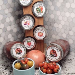 May include: A collection of six glass jars with labels, filled with various homemade preserves. The jars are arranged on a wooden rack. The labels feature hand-drawn illustrations of the ingredients, such as tomatoes, figs, and peppers. The jars are surrounded by small bowls of red tomatoes.