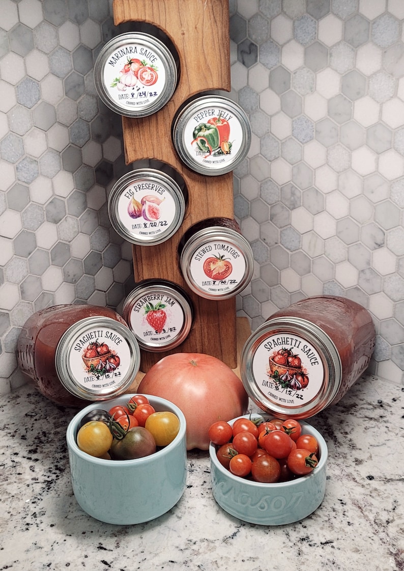 May include: A collection of six glass jars with labels, filled with various homemade preserves. The jars are arranged on a wooden rack. The labels feature hand-drawn illustrations of the ingredients, such as tomatoes, figs, and peppers. The jars are surrounded by small bowls of red tomatoes.