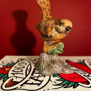 May include: A ceramic bird figurine with brown and tan feathers, perched on a gray tree stump with green leaves. The word "BROAD" is printed on the bird's chest. The figurine is set against a red background.