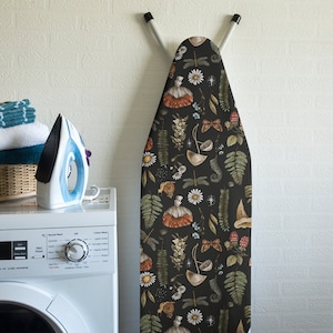 Op de afbeelding: Een zwarte strijkplankhoes met een bloemenpatroon met paddenstoelen, bladeren en vlinders. De hoes hangt aan een muur.