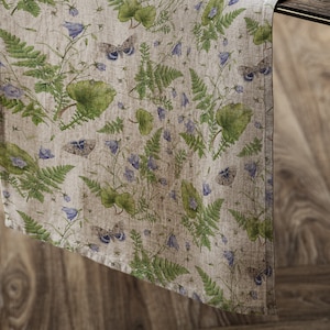Op de afbeelding: Een beige linnen tafelloper met een herhalend patroon van groene varens, blauwe klokjes en vlinders.