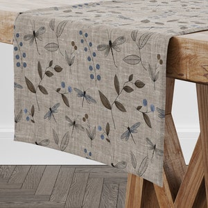 Op de afbeelding: Een beige linnen tafelkleed met een herhalend patroon van blauwe libellen, bruine bladeren en blauwe bessen.