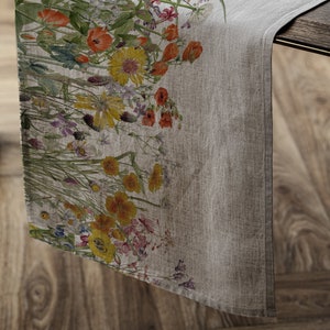 Könnte beinhalten: Ein beigefarbener Tischläufer aus Leinen mit einem Blumendruck, der eine Vielzahl von bunten Blumen zeigt, darunter gelbe, orange, rosa und violette.