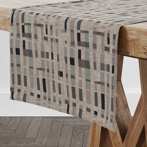 Può includere: Un runner da tavolo beige con un motivo geometrico di rettangoli neri, grigi e marroni. Il runner è realizzato in un tessuto simile al lino e ha una texture sottile.