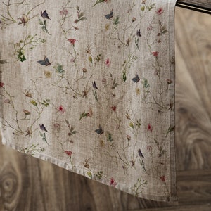 以下が含まれることがあります： ベージュのリネン製のテーブルランナーで、花柄と蝶のプリントが施されています。プリントには、ピンク、ブルー、グリーンの小さな花と蝶が描かれています。