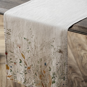 May include: A beige linen table runner with a floral print featuring delicate wildflowers in shades of green, brown, yellow, and blue. The runner is on a wooden table.