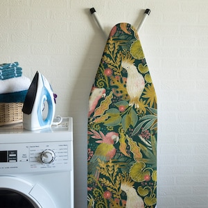 Könnte beinhalten: Ein grüner Bügelbrettbezug mit tropischem Muster mit weißen Kakadus, rosa und grünen Papageien und gelbem und grünem Laub.