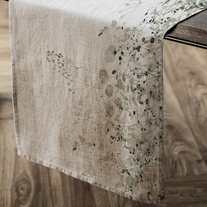 May include: A beige linen table runner with a green floral pattern. The runner has a subtle, faded look, giving it a vintage feel.